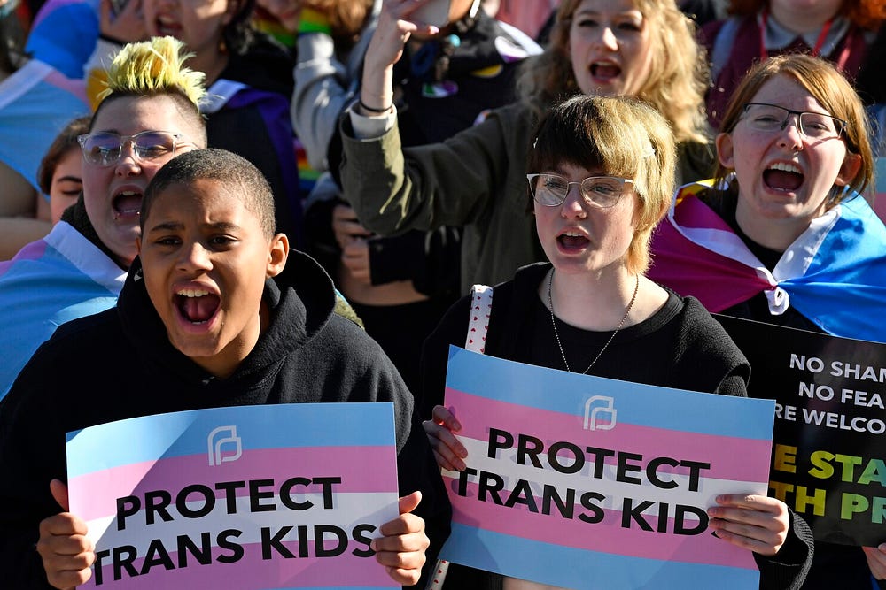 A group of diverse individuals passionately protesting with signs that read 'Protect Trans Kids' and displaying pride flags, advocating for trans rights.